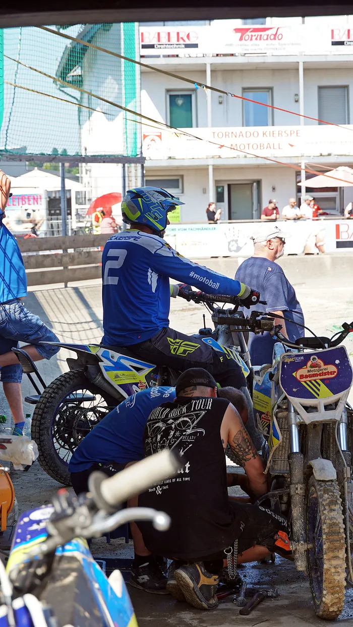Ein Motoballspieler in blauem Rennanzug sitzt auf seinem Motorrad, während ein Mechaniker an der Maschine arbeitet. Im Hintergrund sind weitere Spieler und Zuschauer zu sehen.