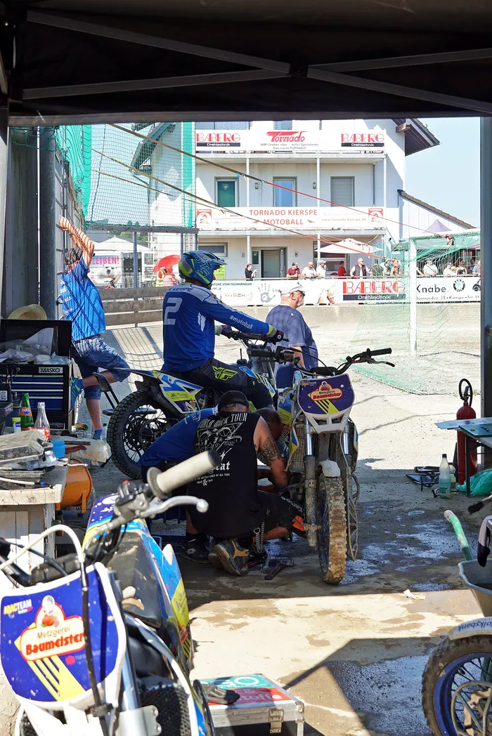 Motoballspieler in voller Ausrüstung sitzen auf ihren Motorrädern in einer Werkshalle. Im Hintergrund ist ein Stadion mit Zuschauern und Werbebannern zu sehen.