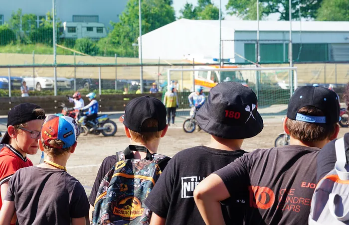 Zuschauer stehen mit dem Rücken zum Betrachter da und beobachten ein Motoball-Rennen auf einer sandigen Bahn. Einige tragen Basecaps, einer einen Rucksack mit Dinosaurier-Motiv.