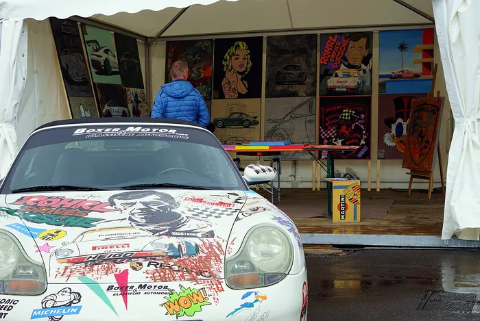 Weißer Porsche mit farbenfroher Lackierung steht vor mehreren Rennsportplakaten in einem Zelt.