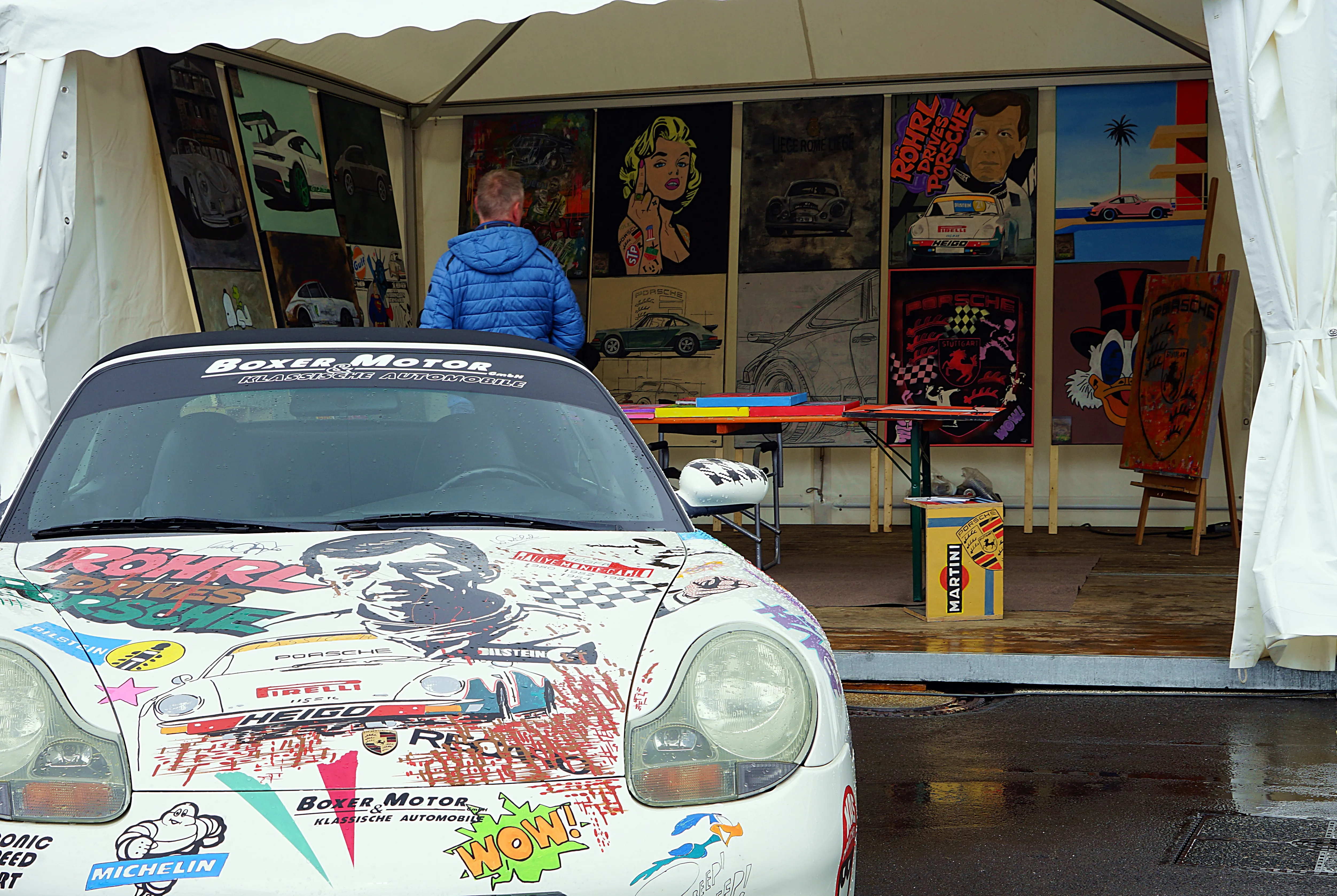 Weißer Porsche mit farbenfroher Lackierung steht vor mehreren Rennsportplakaten in einem Zelt.