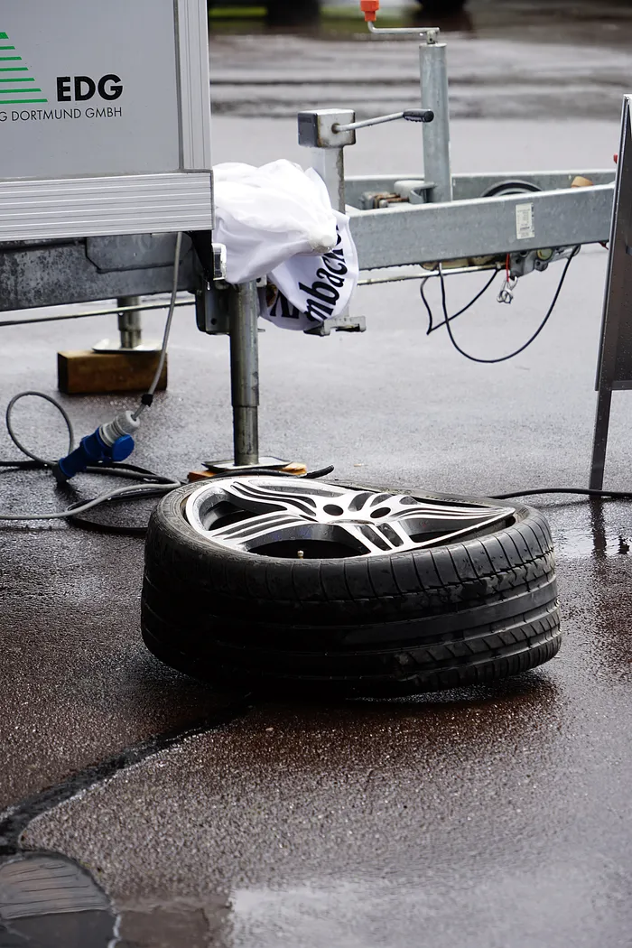 Ein Autoreifen mit Fuchsfelge liegt auf nassem Asphalt vor einem Anhänger mit dem Aufdruck EDG Dortmund GmbH.