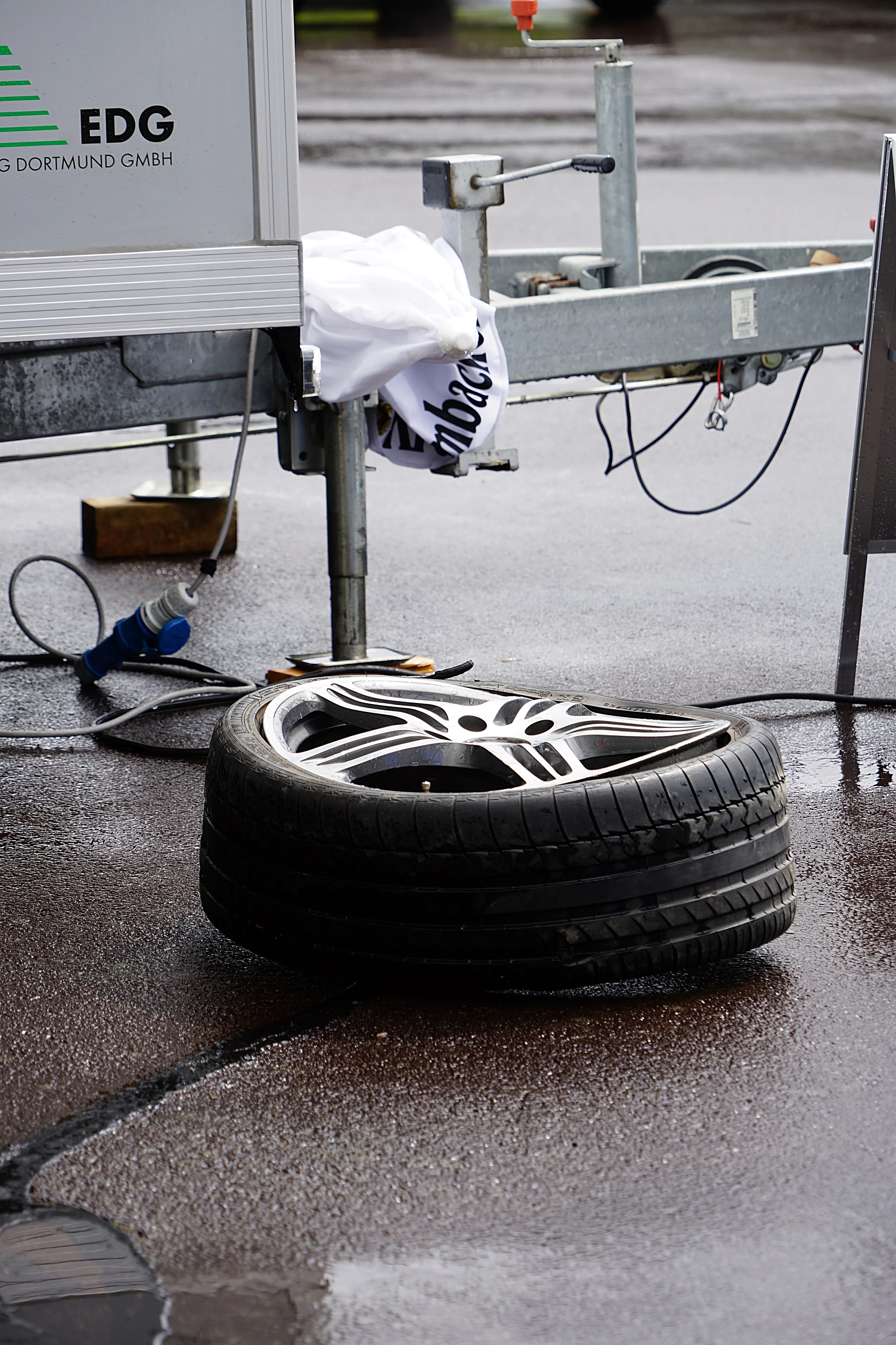 Ein Autoreifen mit Fuchsfelge liegt auf nassem Asphalt vor einem Anhänger mit dem Aufdruck EDG Dortmund GmbH.