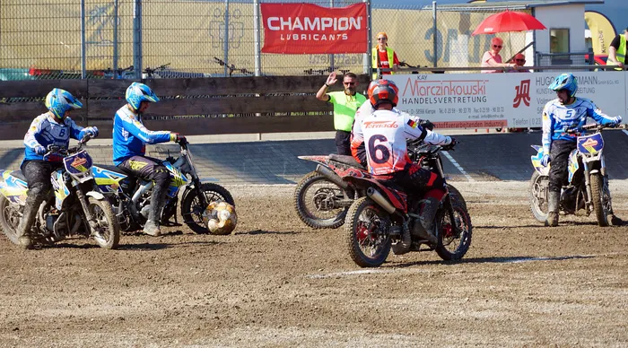 Motoballspieler auf Motorrädern starten in ein Rennen, ein Schiedsrichter mit hochgehobener Hand signalisiert den Beginn. Im Hintergrund Werbebanner und Zuschauer unter Sonnenschirmen.