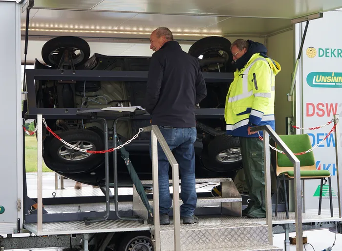 Zwei Männer betrachten einen dunklen Geländewagen auf einem Anhänger, der mit einer Rampe abgelassen wird. Im Hintergrund ein weißer Transporter mit Werbeaufdruck.