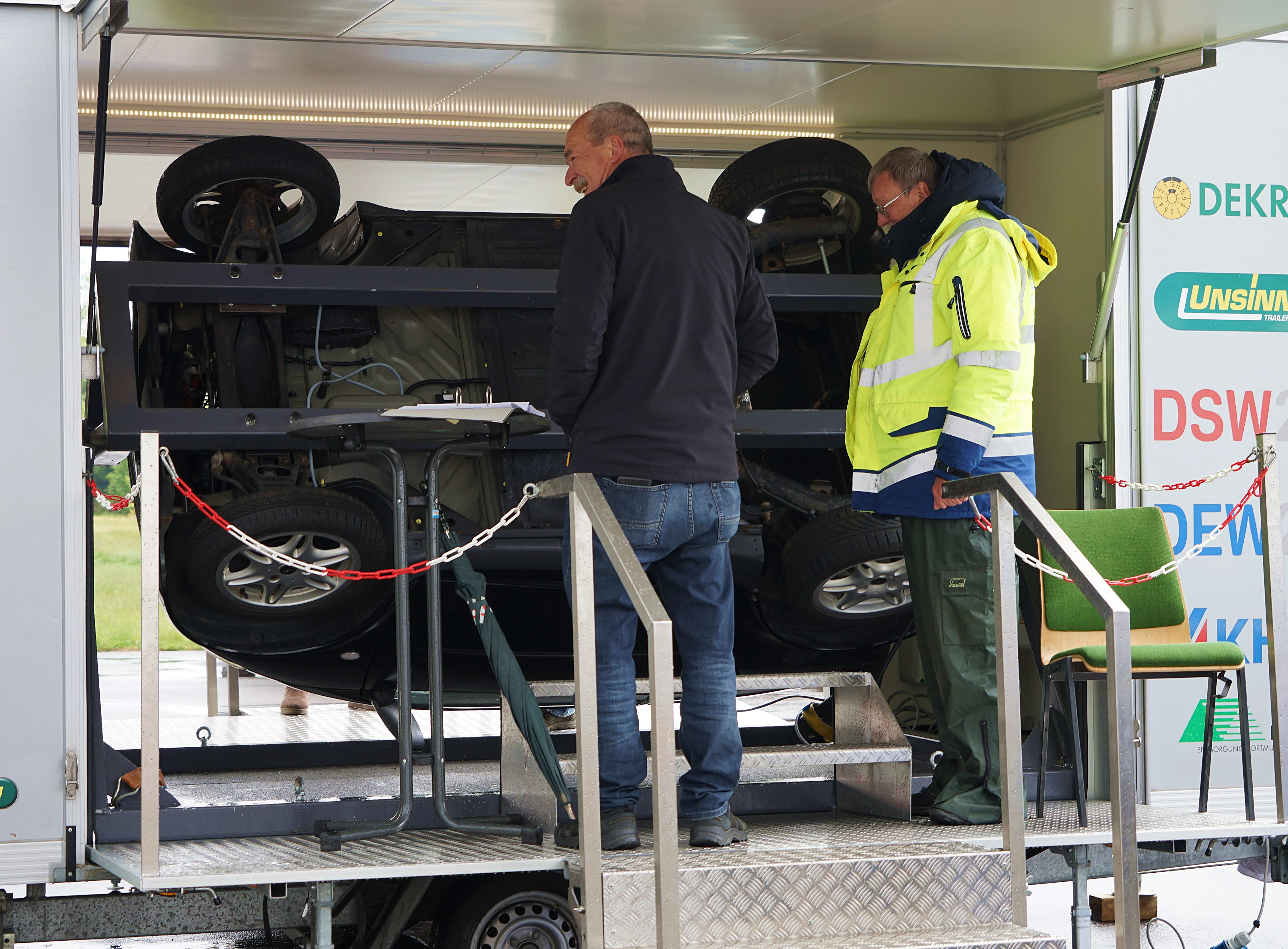 Zwei Männer betrachten einen dunklen Geländewagen auf einem Anhänger, der mit einer Rampe abgelassen wird. Im Hintergrund ein weißer Transporter mit Werbeaufdruck.