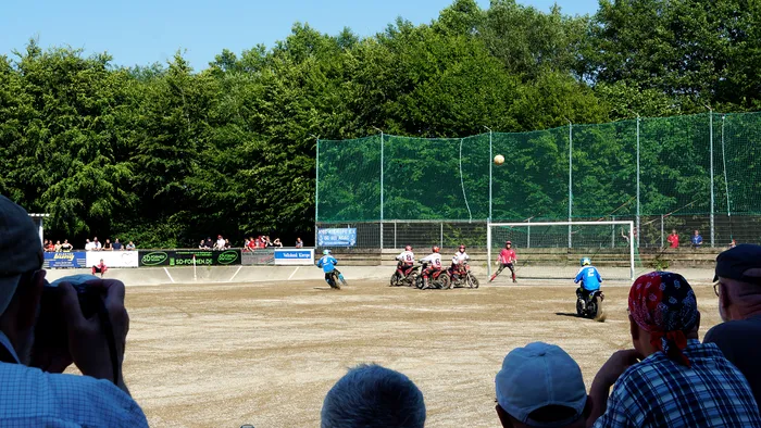 Zwei Motorradfahrer kämpfen um einen Ball auf einem sandigen Spielfeld, während im Hintergrund ein Tor mit Netzen steht und Zuschauer zusehen.