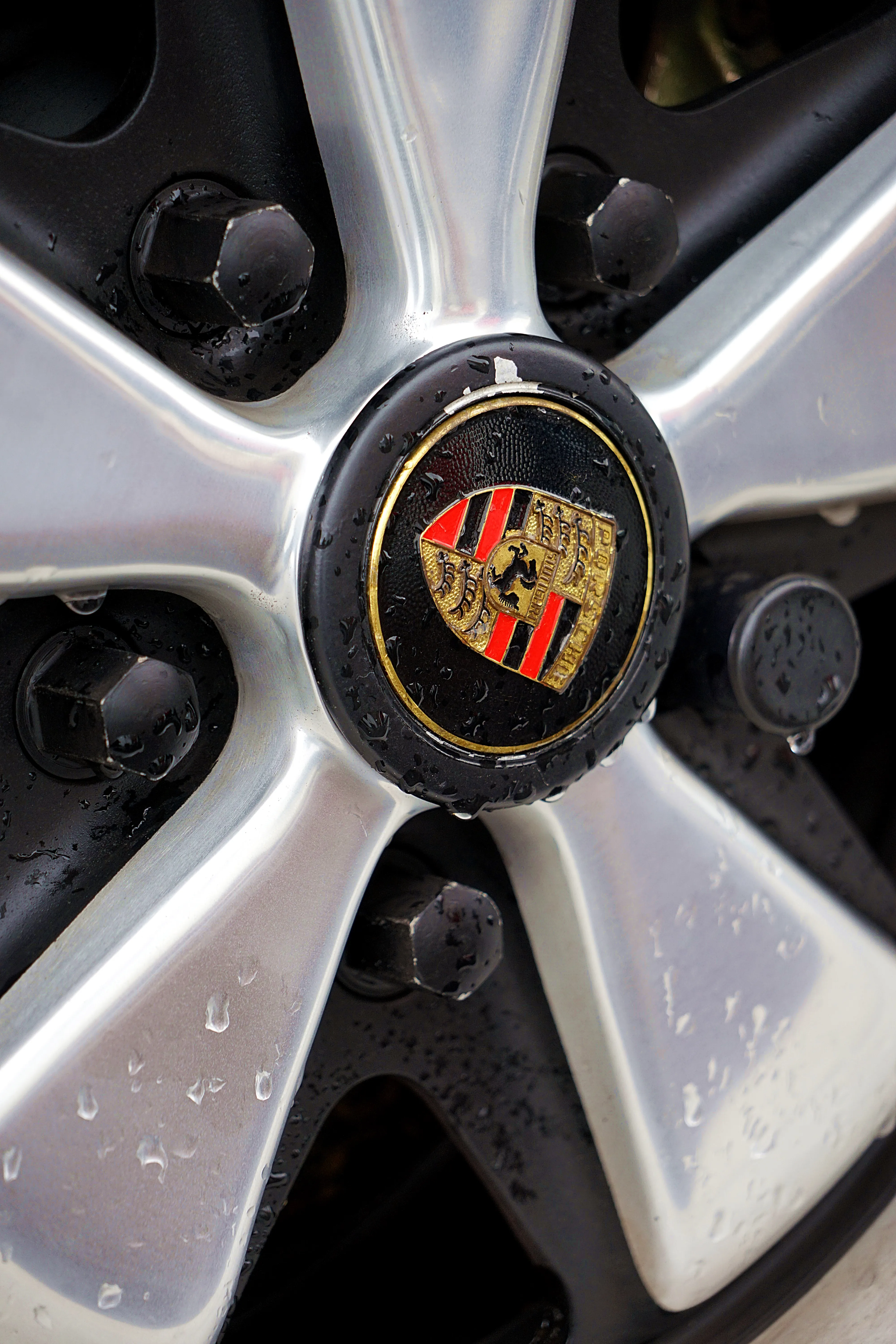 Porsche-Emblem in der Nabe einer silbernen Fuchsfelge, umgeben von Tropfen.