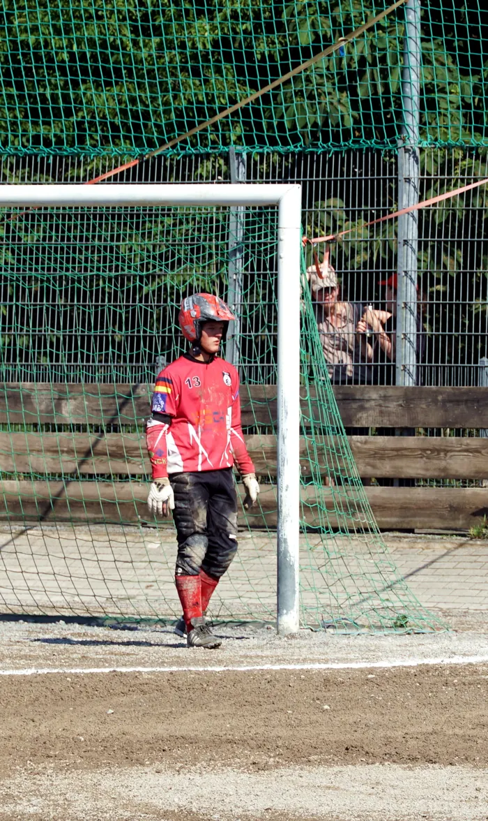 Ein Torwart in roter Ausrüstung und Helm steht vor einem Tor auf einem Feld. Im Hintergrund ist ein Zaun und ein Zuschauer zu sehen.
