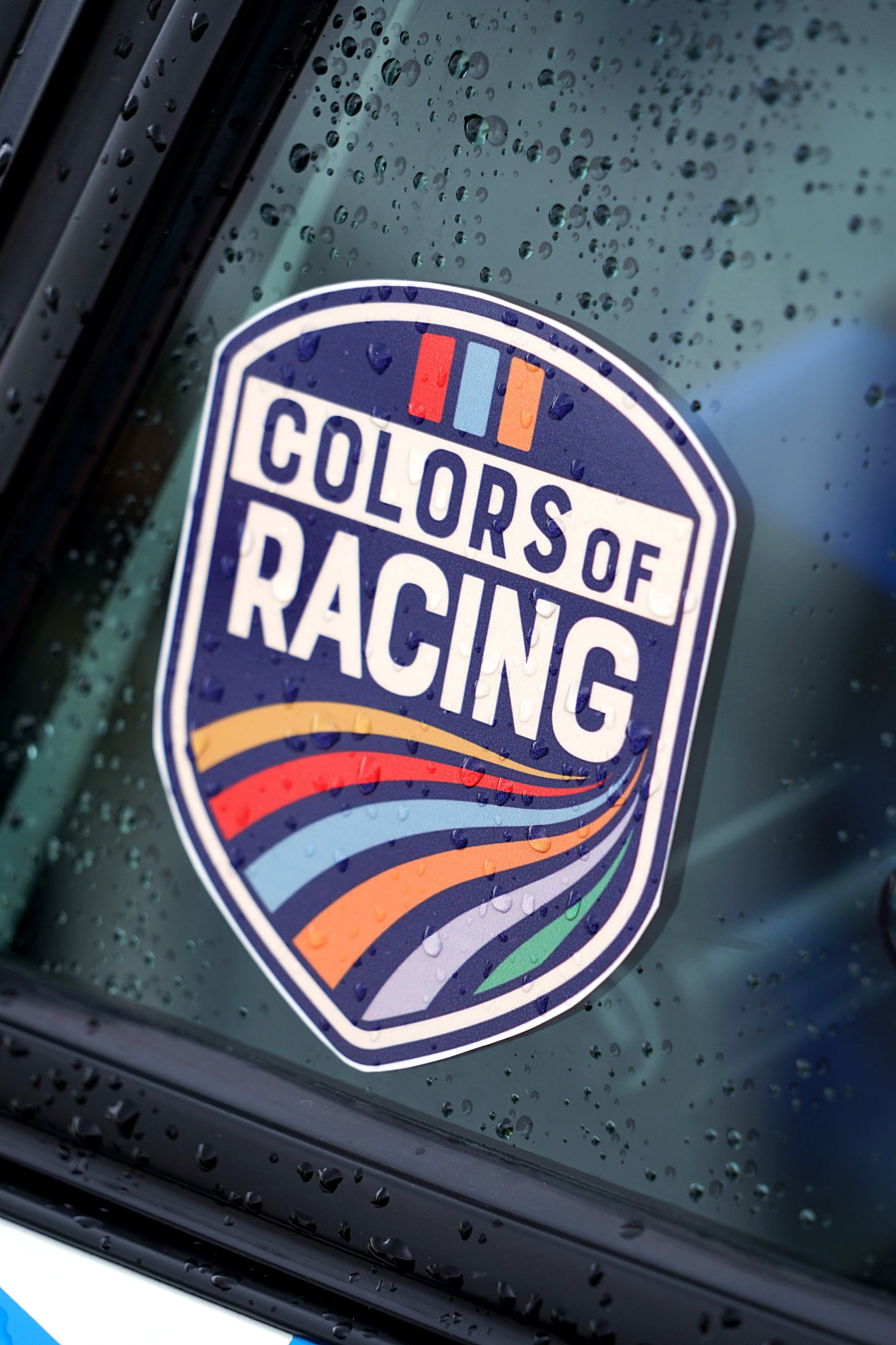 Aufkleber mit der Aufschrift „Colors of Racing“ in den Farben Blau, Orange, Gelb und Rot auf einer Autoscheibe mit Regentropfen.