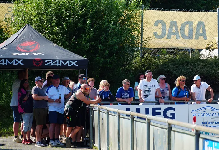 Eine Gruppe von Zuschauern steht hinter einer Metallbarriere und beobachtet ein Motoball-Spiel. Im Hintergrund sind Werbebanner und ein Zelt mit der Aufschrift „24MX“ zu sehen.