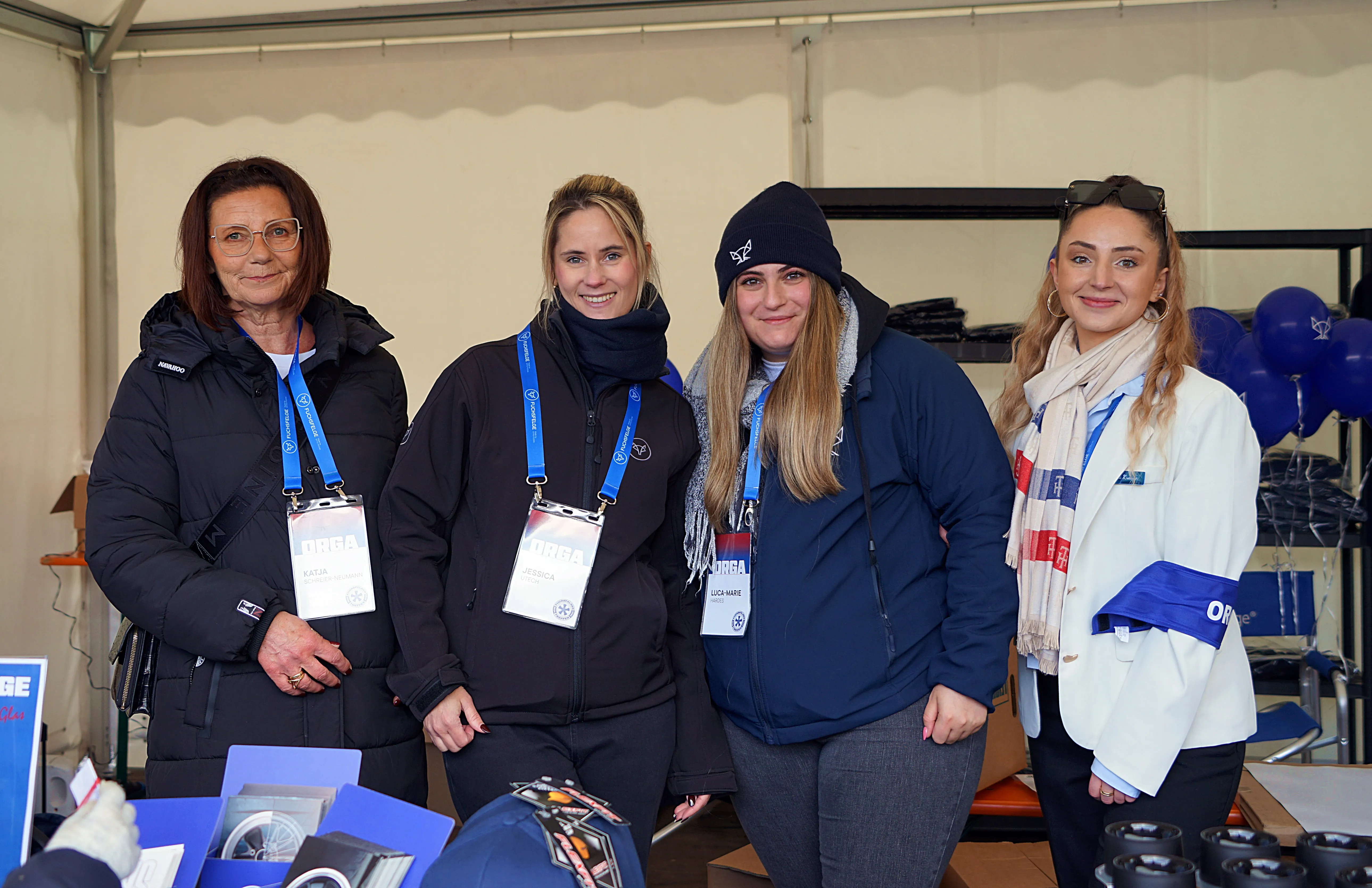 Vier Frauen mit ORGA-Ausweisen stehen nebeneinander vor einem Stand. Sie tragen Winterkleidung in Blau, Schwarz und Weiß.