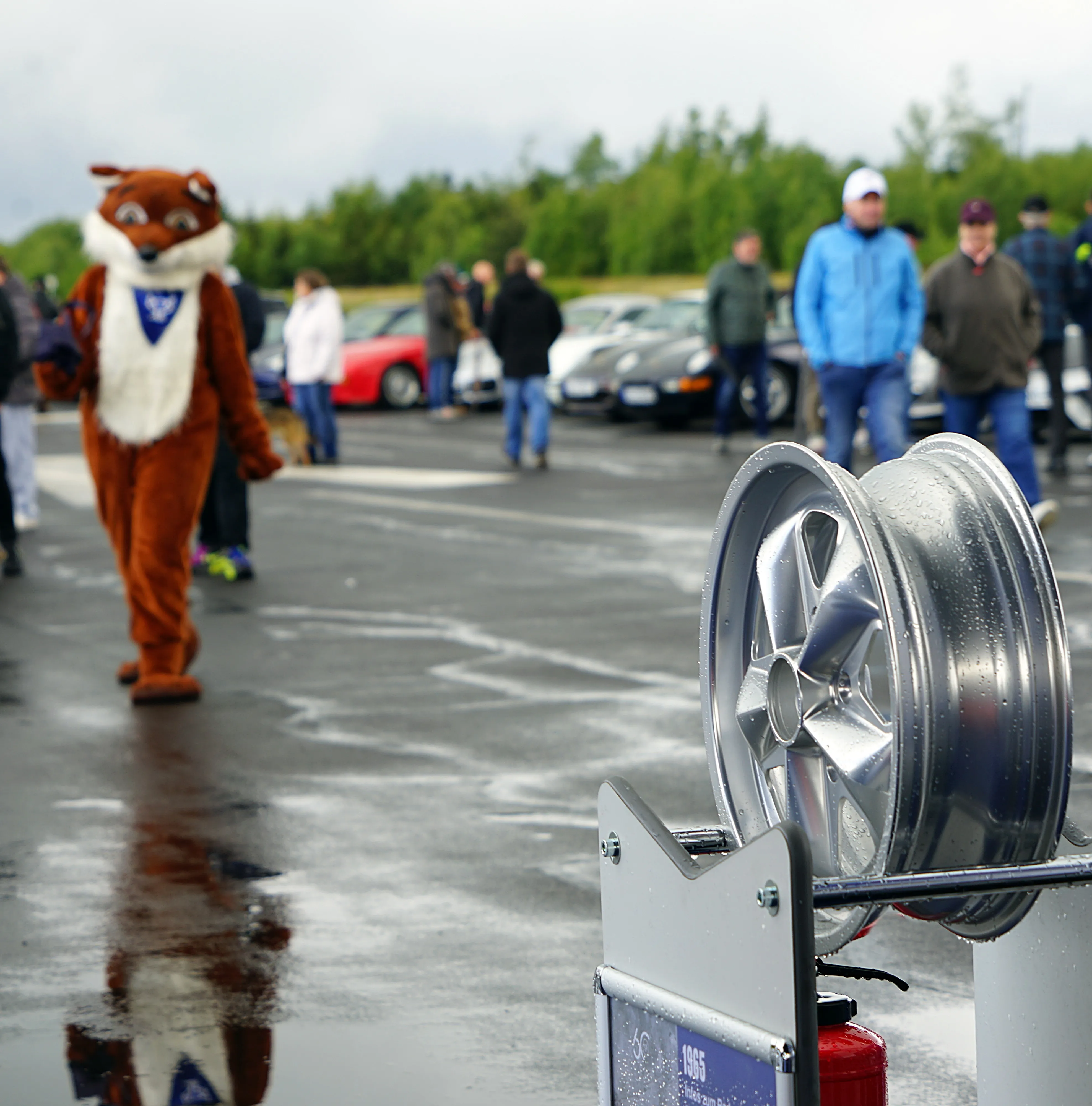 Silberne Fuchsfelge im Vordergrund, im Hintergrund Menschen und Autos auf einem Parkplatz bei bewölktem Himmel.