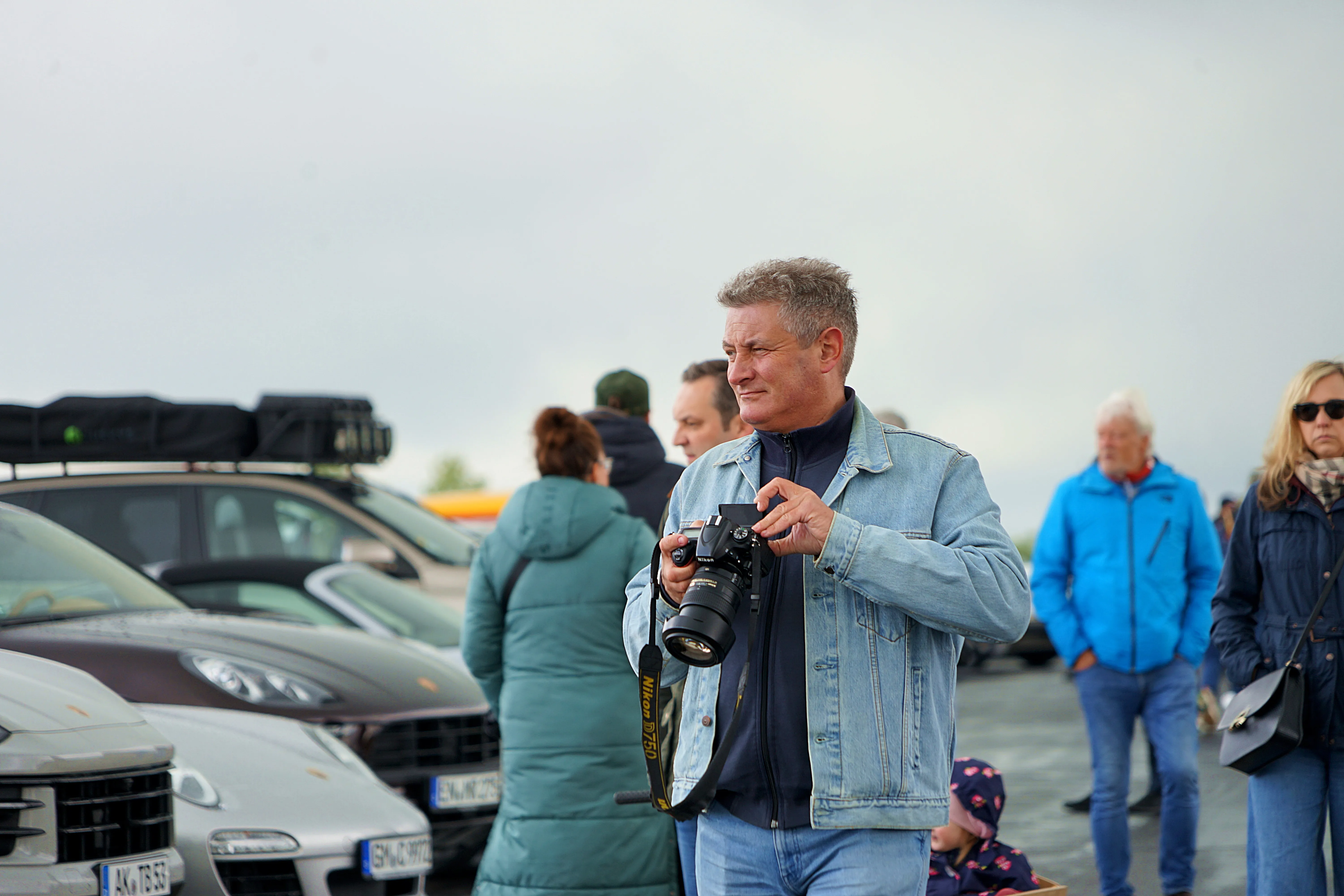 Ein Mann mit grauen Haaren fotografiert mit einer Spiegelreflexkamera auf einem Treffen, umgeben von anderen Besuchern und geparkten Autos.