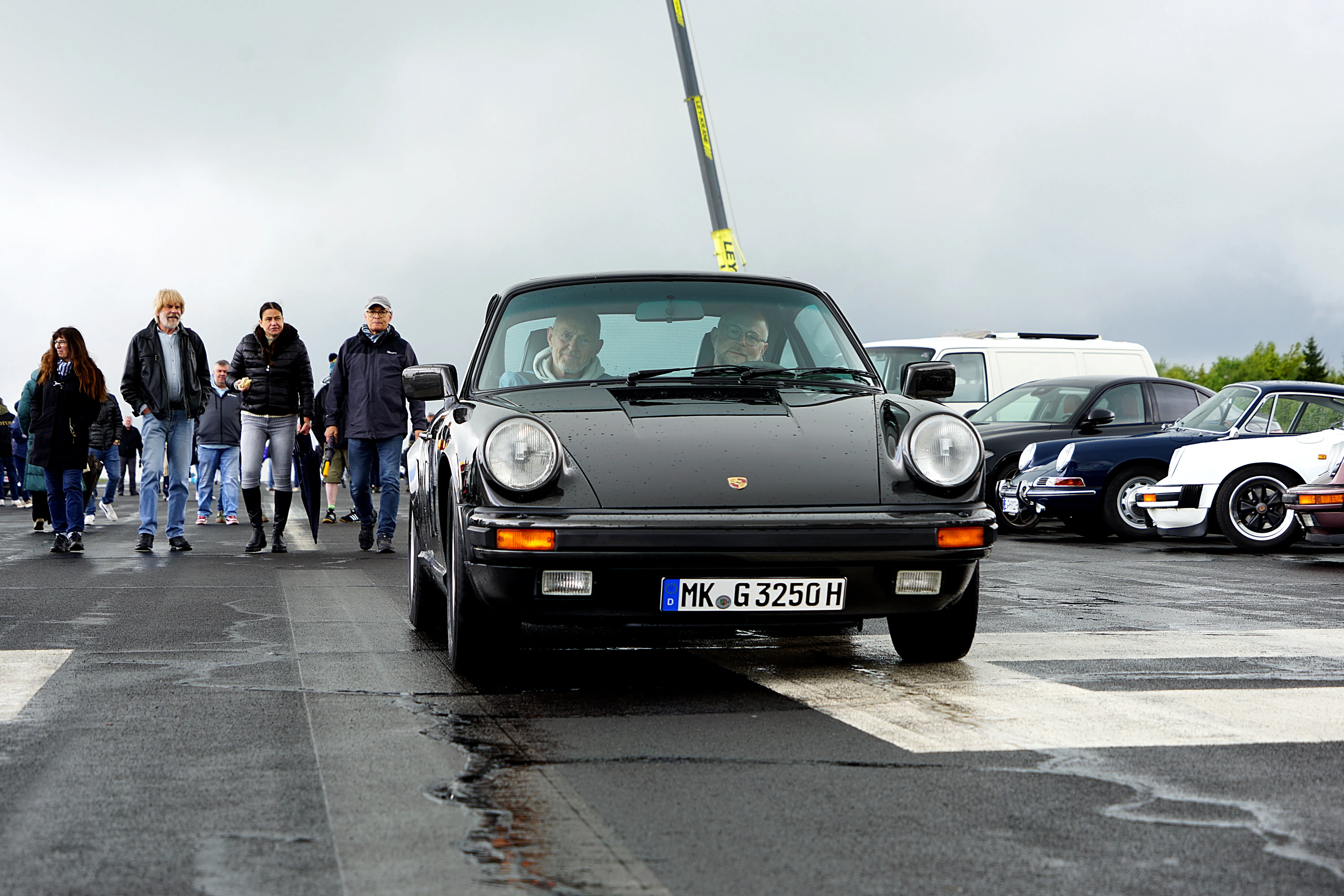 Schwarzer Porsche 911 fährt auf einer nassen Straße vor einer Gruppe von Zuschauern, im Hintergrund weitere Oldtimer.