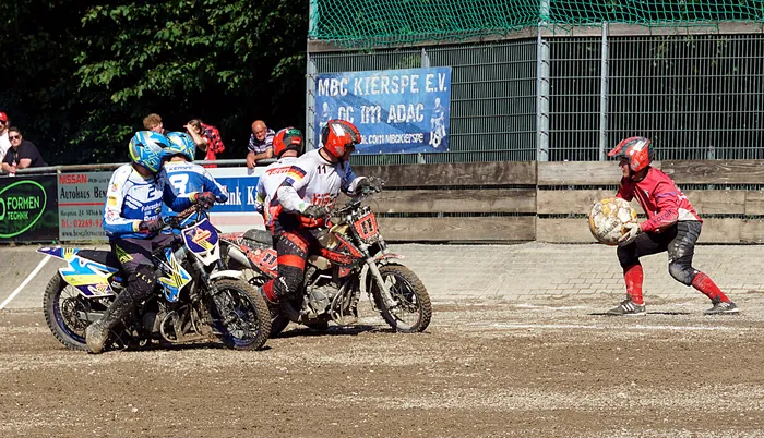 Mehrere Motoballspieler auf Motorrädern kämpfen um den Ball gegen einen Torwart. Im Hintergrund ist ein Banner mit der Aufschrift 'MBC Kierspe e.V.' zu sehen.