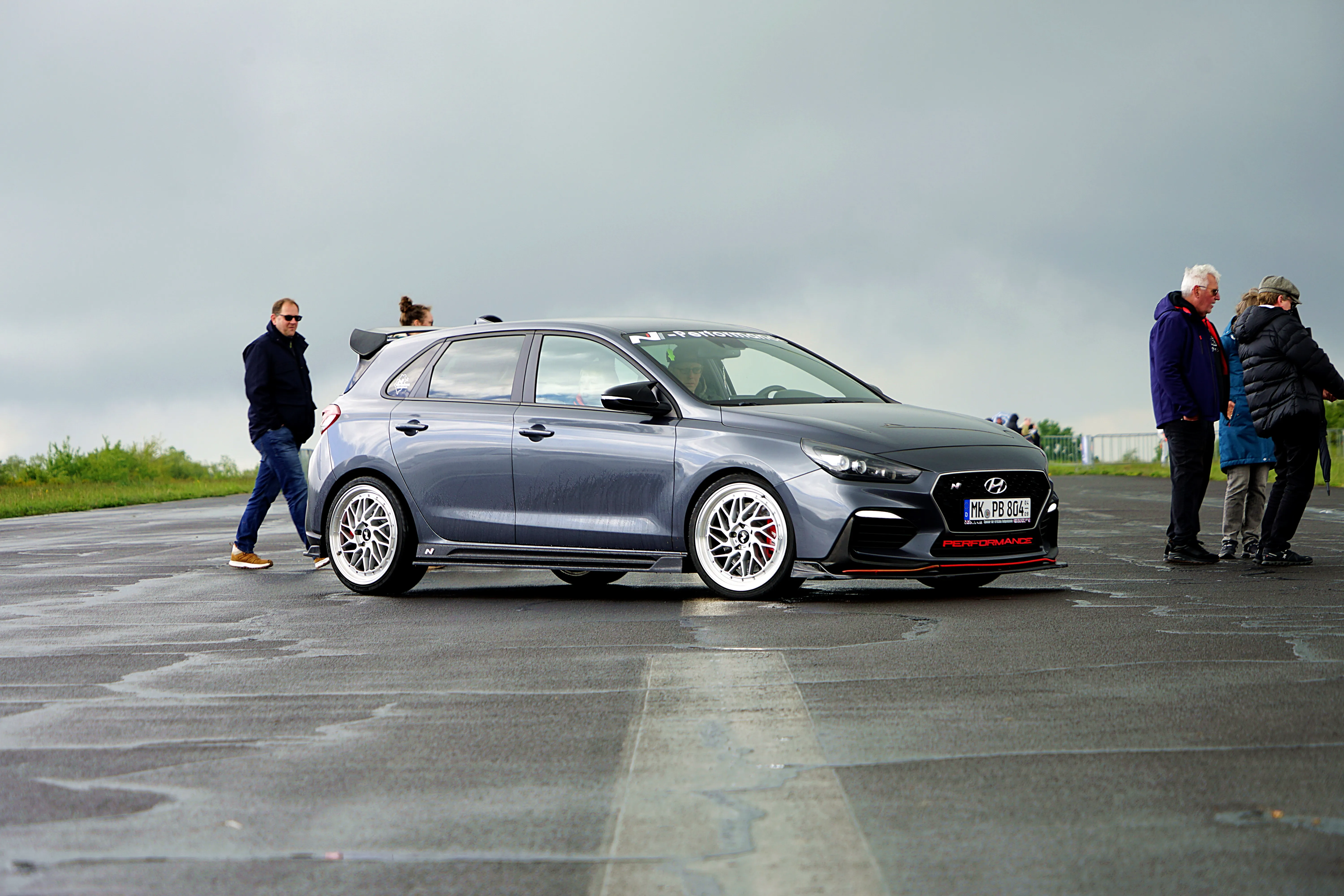Grauer Hyundai i30 N mit Tuning-Elementen und auffälligen Felgen steht auf einem Asphaltplatz, umgeben von Zuschauern.