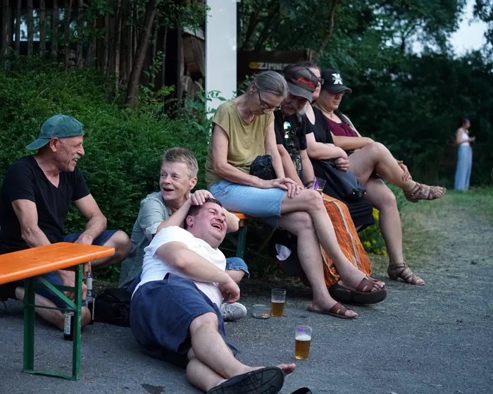 Eine Gruppe von Menschen sitzt und lehnt vor einem Gebäude im Freien. Ein Mann liegt lachend auf dem Boden, während eine Frau ihm die Haare streichelt. Biergläser stehen auf einem Tisch und dem Boden.