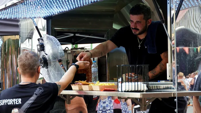 Ein Mann mit Tattoos bereitet Speisen in einem Foodtruck zu und reicht sie einem Kunden über den Tresen. Im Hintergrund sind weitere Personen und ein Ventilator zu sehen.