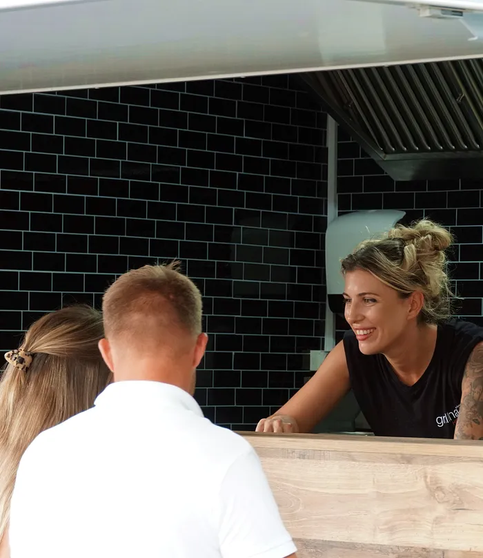 Eine Frau mit blonden Haaren und Tätowierungen lächelt über einen Tresen hinweg zu einer Person mit kurzen blonden Haaren. Im Hintergrund eine schwarze Fliesenwand und eine Dunstabzugshaube.