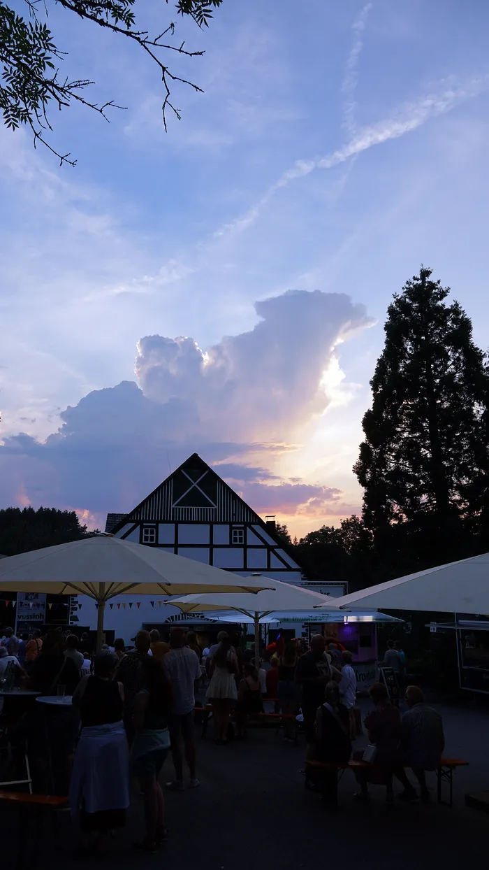 Ein Abend am See: Menschen sitzen an Biertischbänken unter Sonnenschirmen vor einem traditionellen Fachwerkhaus, während die Sonne hinter Wolken untergeht.