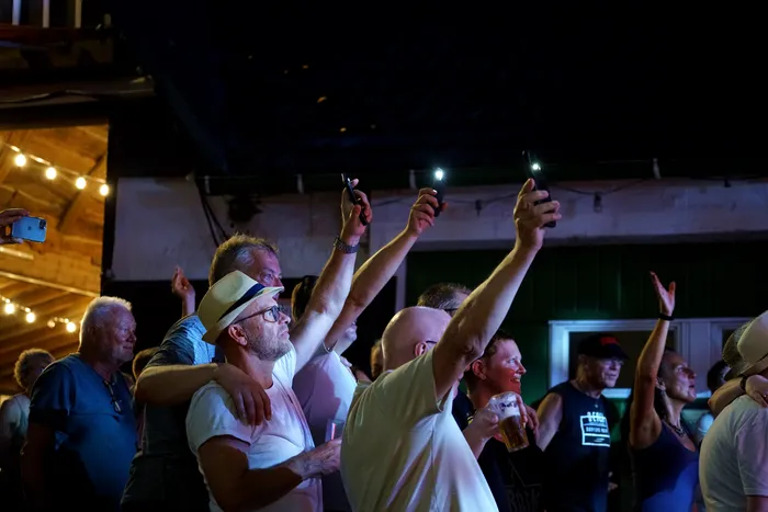 Eine Gruppe von Männern steht eng beieinander und filmt oder fotografiert mit ihren Smartphones etwas außerhalb des Bildausschnitts. Einige halten Biergläser in der Hand, die Stimmung wirkt ausgelassen. Lichterketten beleuchten die Szene.