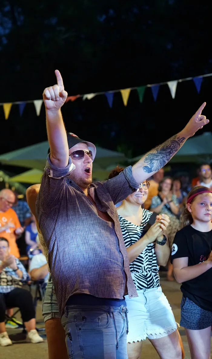 Ein Mann mit Tätowierungen auf den Armen hebt bei einem Open-Air-Event beide Hände in die Luft, während im Hintergrund Menschen sitzen und stehen. Girlanden sind über die Szene gespannt.