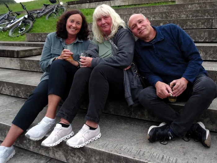 Drei lächelnde Menschen sitzen auf einer Betontreppe und halten jeweils ein Glas in der Hand. Im Hintergrund sind Fahrräder und weitere Treppenstufen zu sehen.