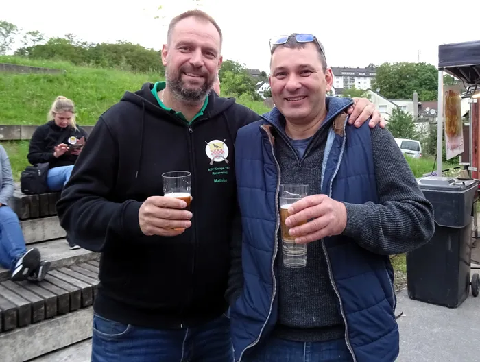 Zwei lächelnde Männer halten jeweils ein Glas Bier in der Hand und stehen vor einer Grünfläche. Im Hintergrund sind weitere Personen und ein Gebäude zu sehen.