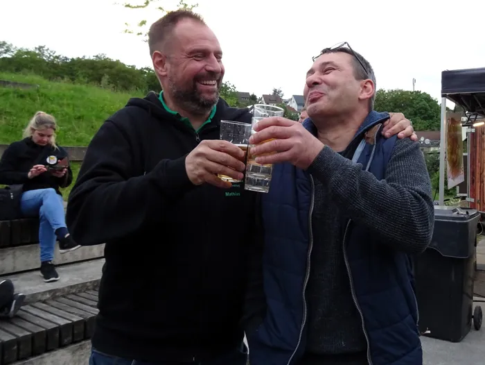 Zwei Männer stoßen mit Biergläsern an und lächeln in die Kamera. Im Hintergrund sitzt eine Frau auf einer Bank und benutzt ihr Smartphone.