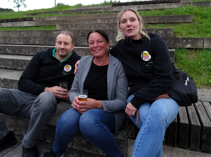 Drei lächelnde Menschen sitzen auf einer Stufe vor einer Treppe im Freien. Die Frau in der Mitte hält ein Bierglas, die anderen haben Getränke neben sich stehen. Alle tragen Jacken mit einem Logo darauf.