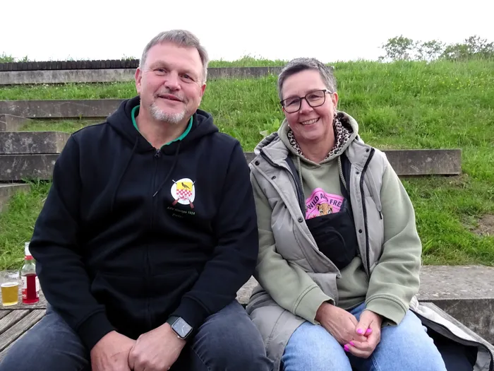 Ein Mann und eine Frau sitzen lächelnd nebeneinander auf einer Holzbank. Der Mann trägt einen schwarzen Kapuzenpullover mit einem Wappen darauf, die Frau eine Weste über einem pinkfarbenen T-Shirt. Im Hintergrund ist eine grüne Wiese und eine Steinmauer zu sehen.
