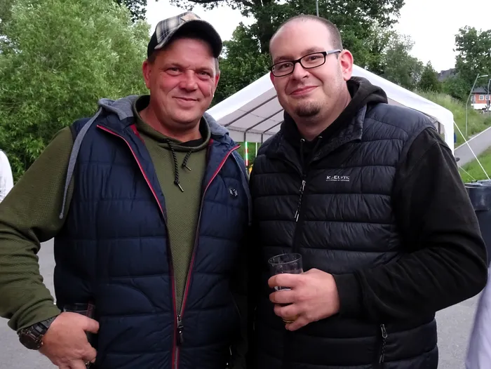 Zwei lächelnde Männer stehen eng nebeneinander im Freien. Der Mann links trägt eine Kappe und eine Weste, der Mann rechts eine Brille und eine dunkle Jacke.
