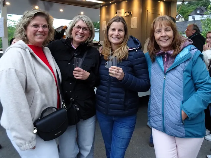 Vier lächelnde Frauen stehen eng zusammen, jede hält ein Glas in der Hand. Im Hintergrund ist ein Foodtruck und weitere Menschen zu sehen.