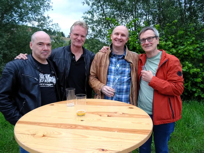 Vier Männer stehen lächelnd um einen runden Holztisch. Auf dem Tisch stehen zwei halbvolle Gläser. Im Hintergrund sind Bäume und Büsche zu sehen.