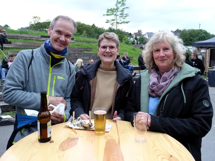 Drei lächelnde Menschen sitzen an einem Holztisch im Freien. Vor ihnen stehen Getränke und ein Teller mit Meeresfrüchten. Im Hintergrund sind weitere Menschen und ein Zelt zu sehen.