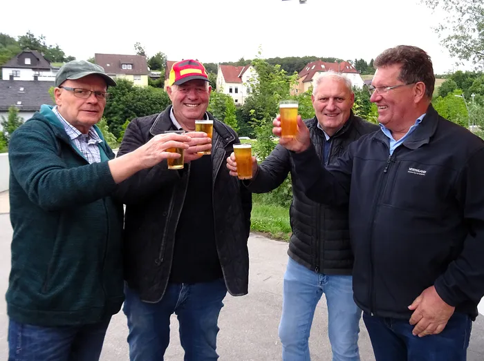 Vier Männer stehen im Freien zusammen und stoßen mit Biergläsern an. Sie lächeln und scheinen eine gute Zeit zu haben. Im Hintergrund sind Häuser und Bäume zu sehen.