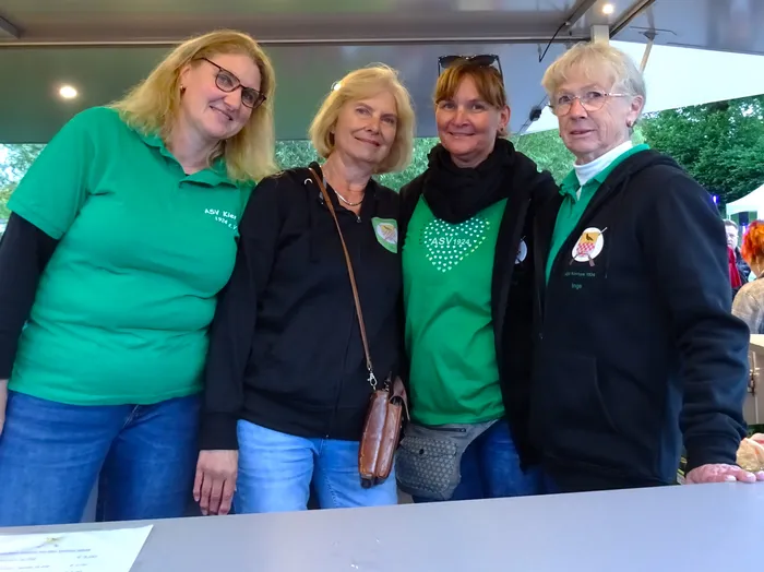 Vier Frauen stehen lächelnd vor einem Stand, vermutlich auf einem Volksfest oder einer ähnlichen Veranstaltung. Sie tragen grüne oder schwarze Kleidung mit dem Logo 