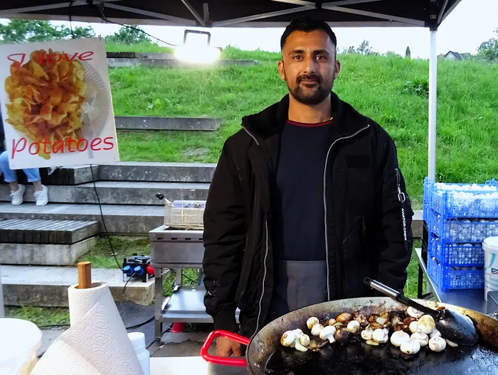 Mann mit dunkler Jacke steht an einem Imbissstand mit Pommes und Bratpfanne vor sich. Im Hintergrund ein Werbeplakat für Pommes und eine grüne Wiese.