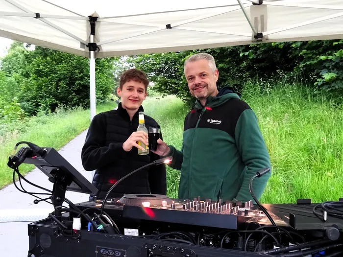 Zwei Männer stehen unter einem Zelt neben einer DJ-Anlage, einer hält eine Flasche. Im Hintergrund ist eine grüne Wiese zu sehen.