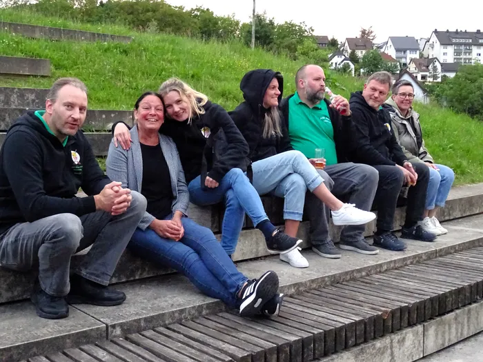 Eine Gruppe von sieben Erwachsenen sitzt lächelnd auf einer Stufe im Freien. Einige halten Getränke in der Hand. Im Hintergrund sind Häuser und Grünflächen zu sehen.