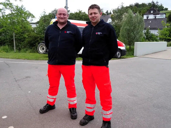 Zwei Männer in Uniformen des Deutschen Roten Kreuzes stehen lächelnd auf einem Asphaltweg. Im Hintergrund ist ein Rettungswagen und ein Wohnhaus zu sehen.