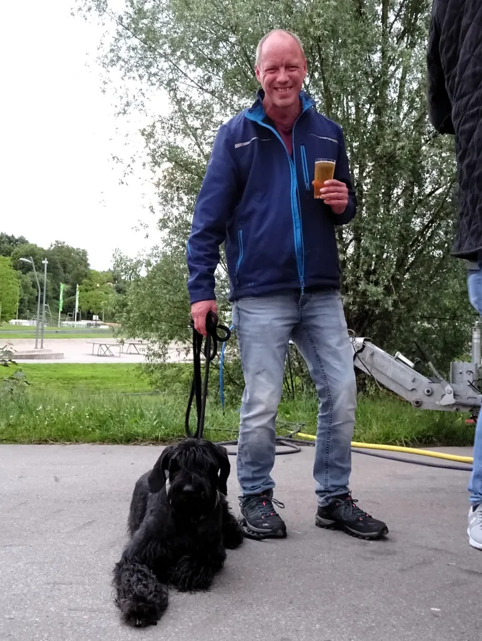 Ein lächelnder Mann mit einem Glas Bier steht neben einem liegenden, schwarzen Cocker Spaniel an einer Leine. Im Hintergrund ist ein Park mit Spielgeräten zu sehen.