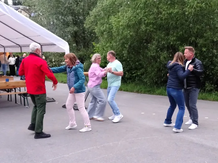 Eine Gruppe von Menschen tanzt im Freien auf einem asphaltierten Platz vor einem Zelt und Bäumen. Die Tänzer sind unterschiedlichen Alters und tragen Freizeitkleidung.