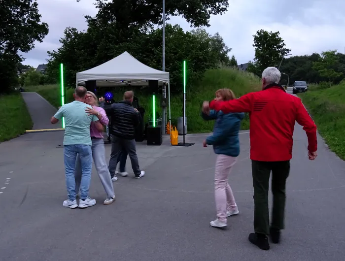 Menschen tanzen im Freien vor einem Zelt, während im Hintergrund Bäume und ein Parkplatz zu sehen sind. Einige tragen Jacken, was auf kühles Wetter hindeutet.