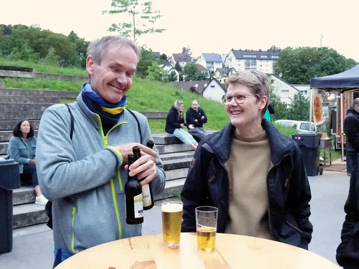 Ein Mann schenkt einer Frau Bier in ein Glas ein, beide lächeln in die Kamera. Im Hintergrund sitzen und stehen weitere Personen auf einem Gelände mit Bäumen und Häusern.