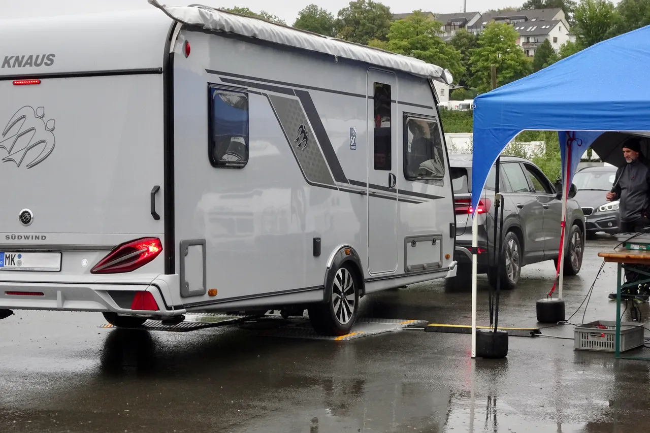 Ein Wohnwagen der Marke Knaus Südwind steht auf einem nassen Parkplatz neben einem blauen Pavillon und einem grauen Auto. Im Hintergrund sind weitere Fahrzeuge und Gebäude zu sehen.