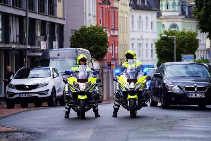 Zwei Polizisten auf gelb-weißen Motorrädern mit Blaulicht fahren auf einer Stadtstraße auf die Kamera zu.