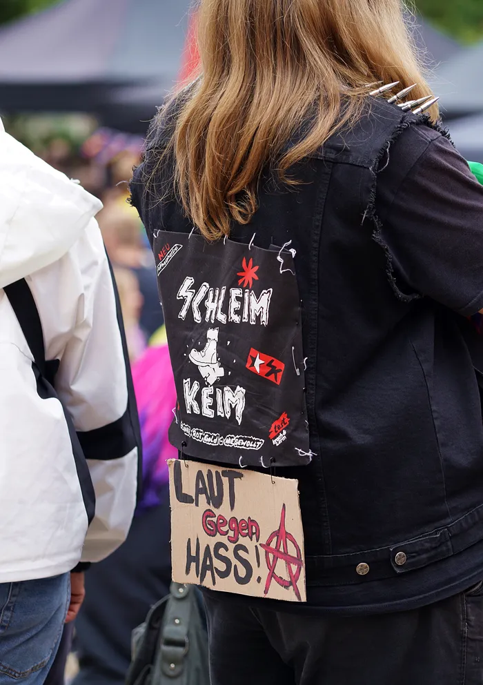 Rückansicht einer Person in einer schwarzen Jacke mit einem Pappschild mit der Aufschrift „LAUT GEGEN HASS!“ und einem Anarchie-Symbol.