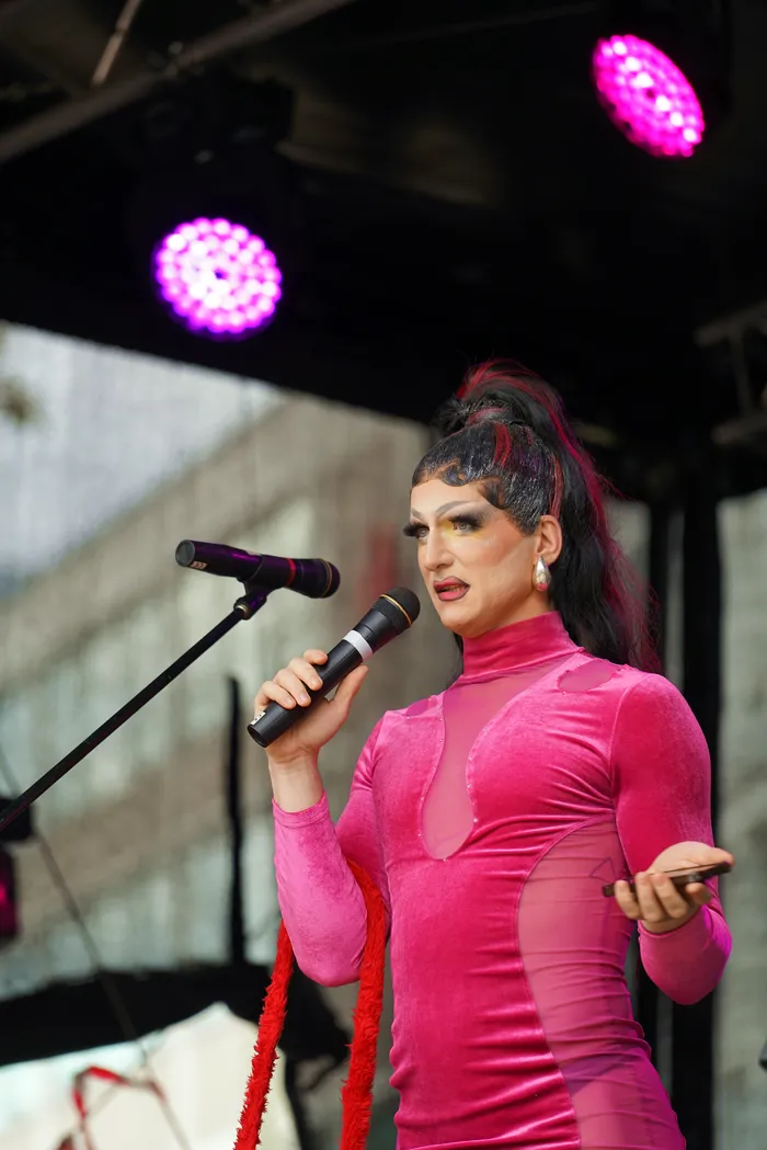 Drag-Performer in einem pinkfarbenen Kleid hält ein Mikrofon und performt auf einer Bühne unter pinken Scheinwerfern.